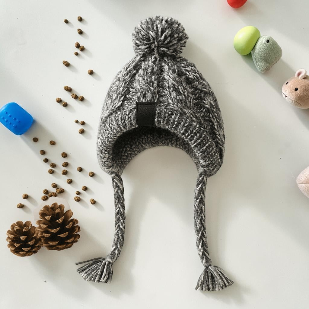 Furvana grey cable-knit dog hat with ear flaps, braided tassels, and a pom-pom on a white background with pinecones.