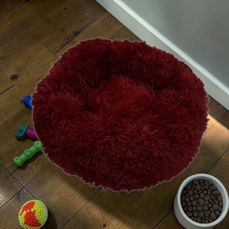 Furvana plush red donut dog bed with raised rim for orthopedic support on a wooden floor with pet toys and food bowl