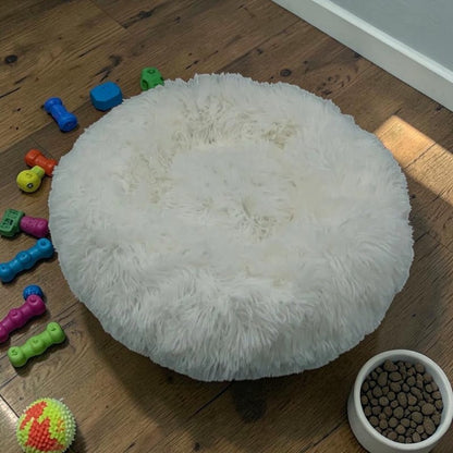 Furvana white plush calming donut bed for dogs and cats with raised rim on a wooden floor with scattered pet toys